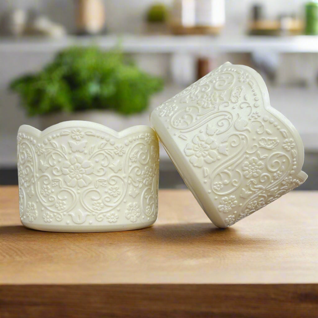 Aalijah Shimmer LUXE Tumbler Boot – A vintage-inspired silicone tumbler boot featuring an intricate 3D floral design with a delicate scalloped edge. Designed to fit most 20-40 oz tumblers, this elegant and upscale tumbler accessory enhances aesthetics while helping protect the base. Shown in a soft ivory hue on a wooden surface with a blurred modern kitchen background and greenery.