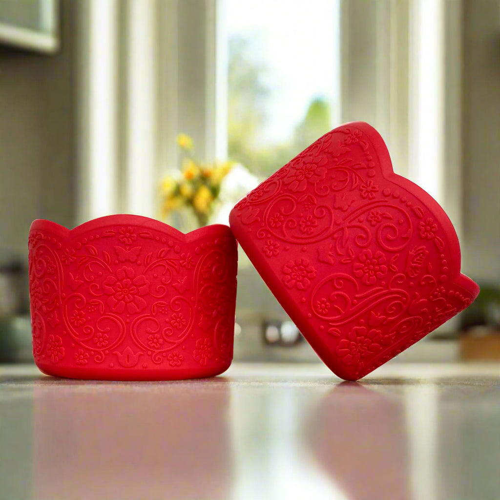 Brielle LUXE Tumbler Boot – A vintage-inspired silicone tumbler boot featuring an intricate 3D floral design with a delicate scalloped edge. Designed to fit most 20-40 oz tumblers, this bold and upscale tumbler accessory enhances aesthetics while helping protect the base. Shown in a striking red hue on a reflective countertop with a blurred kitchen and floral background.