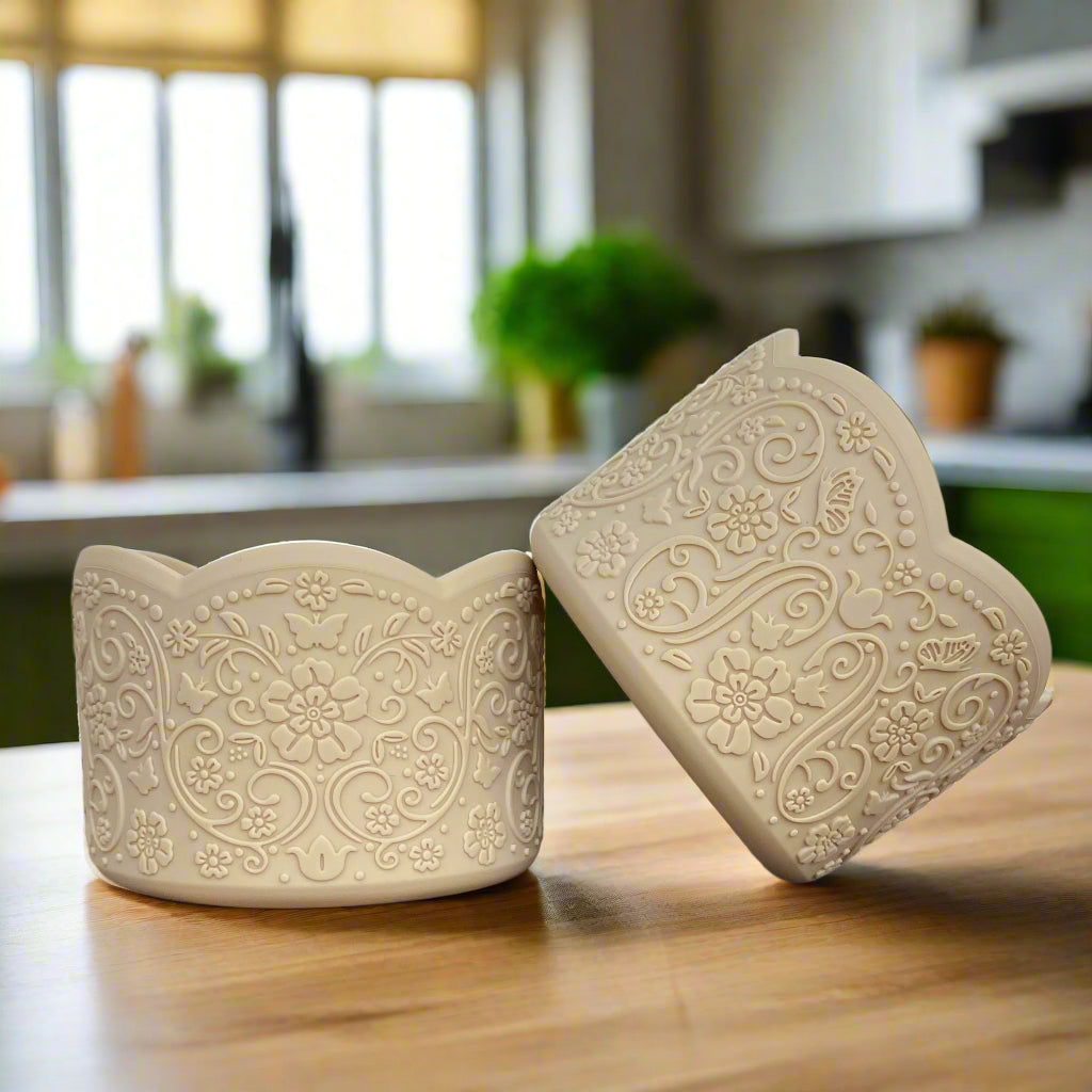 Dawn LUXE Tumbler Boot – A vintage-inspired silicone tumbler boot featuring an intricate 3D floral design with a delicate scalloped edge. Designed to fit most 20-40 oz tumblers, this elegant and upscale tumbler accessory enhances aesthetics while helping protect the base. Shown in a soft neutral beige hue on a wooden surface with a blurred cozy kitchen background