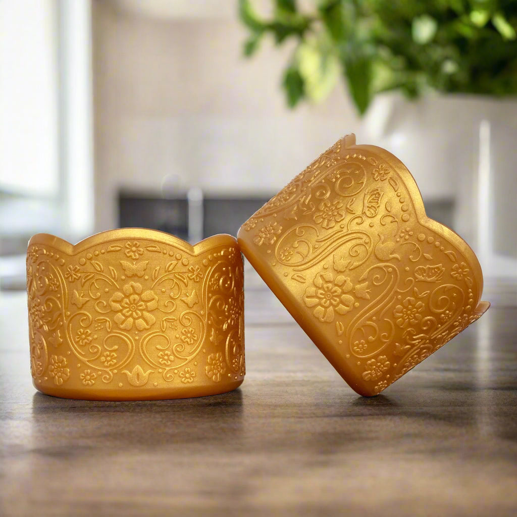 Doris LUXE Tumbler Boot – A vintage-inspired silicone tumbler boot featuring an intricate 3D floral design with a delicate scalloped edge. Designed to fit most 20-40 oz tumblers, this elegant and upscale tumbler accessory enhances aesthetics while helping protect the base. Shown in a rich golden hue with a subtle shimmer, placed on a wooden surface with a blurred modern kitchen background