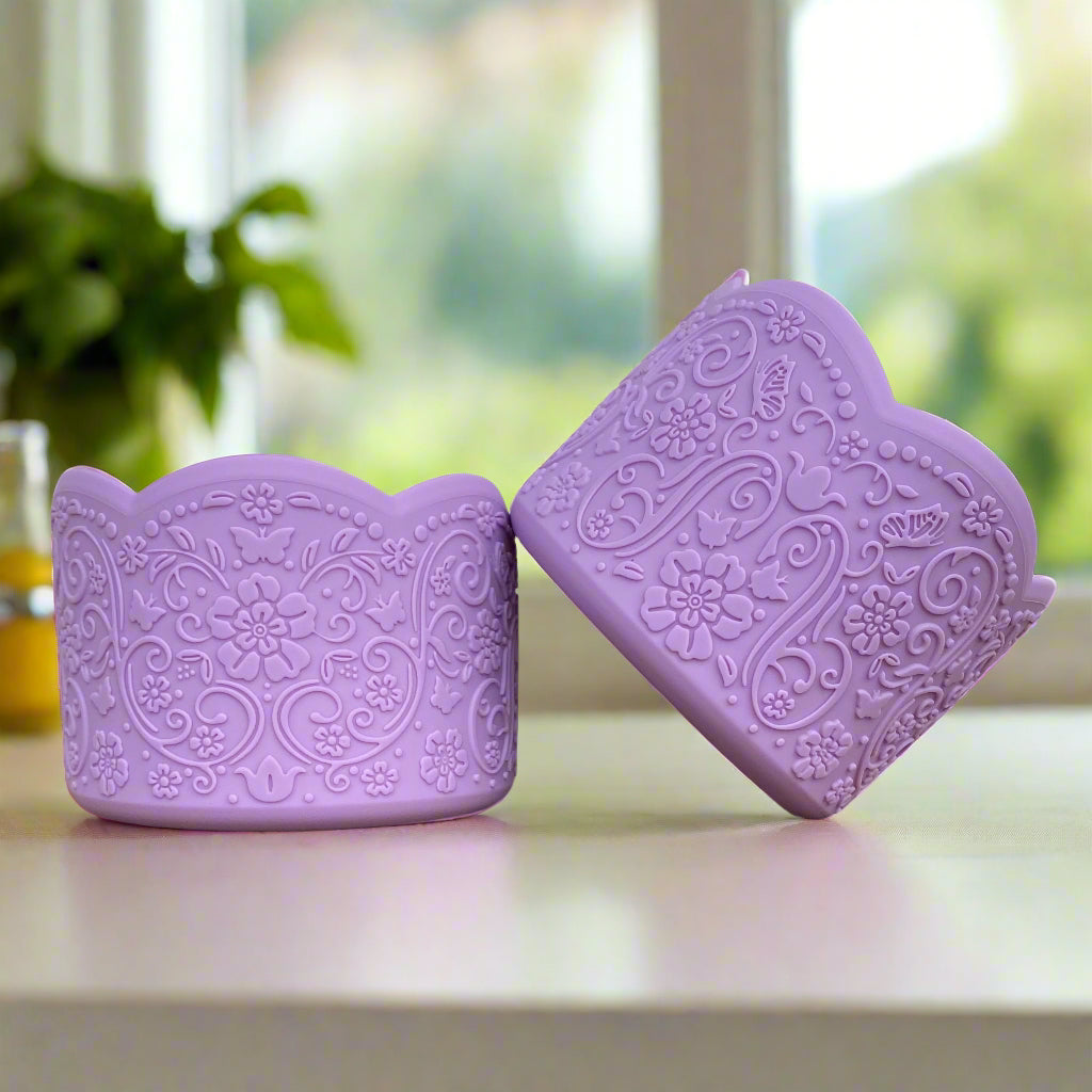 Mia LUXE Tumbler Boot – A vintage-inspired silicone tumbler boot featuring an intricate 3D floral design with a delicate scalloped edge. Designed to fit most 20-40 oz tumblers, this unique and upscale tumbler accessory enhances style while helping protect the base. Shown in a soft lavender hue on a clean, neutral countertop with a blurred sunlit window and greenery in the background