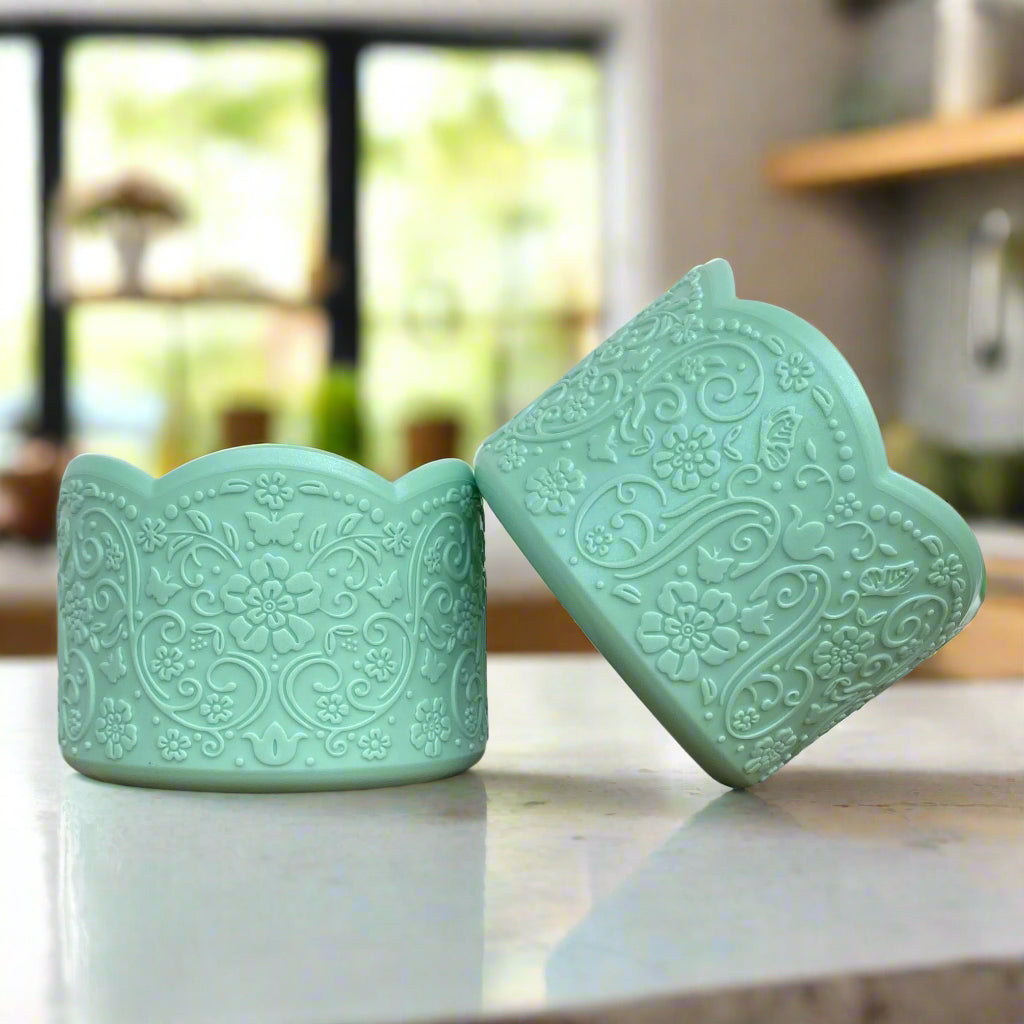 Mindy Shimmer LUXE Tumbler Boot – A vintage-inspired silicone tumbler boot featuring an intricate 3D floral design with a delicate scalloped edge. Designed to fit most 20-40 oz tumblers, this stylish and upscale tumbler accessory adds an elegant touch while helping protect the base. Shown in a soft green pastel hue on a marble countertop with a blurred modern kitchen background.