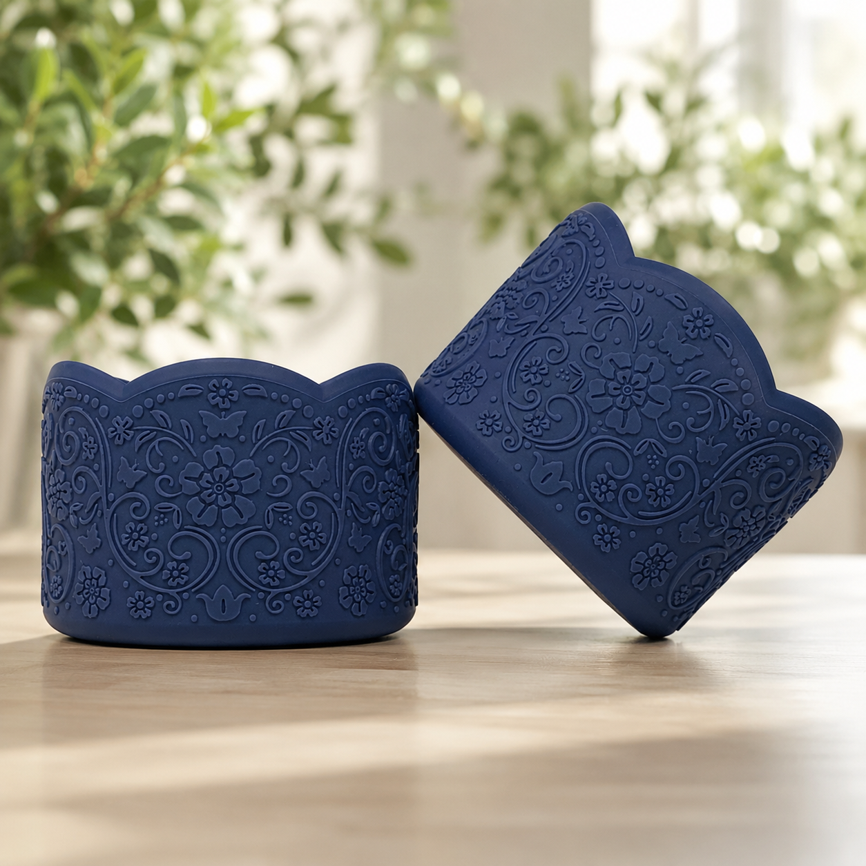 Two navy blue silicone tumbler boots with embossed floral and scroll patterns, displayed on a light wooden surface with a blurred green plant background.