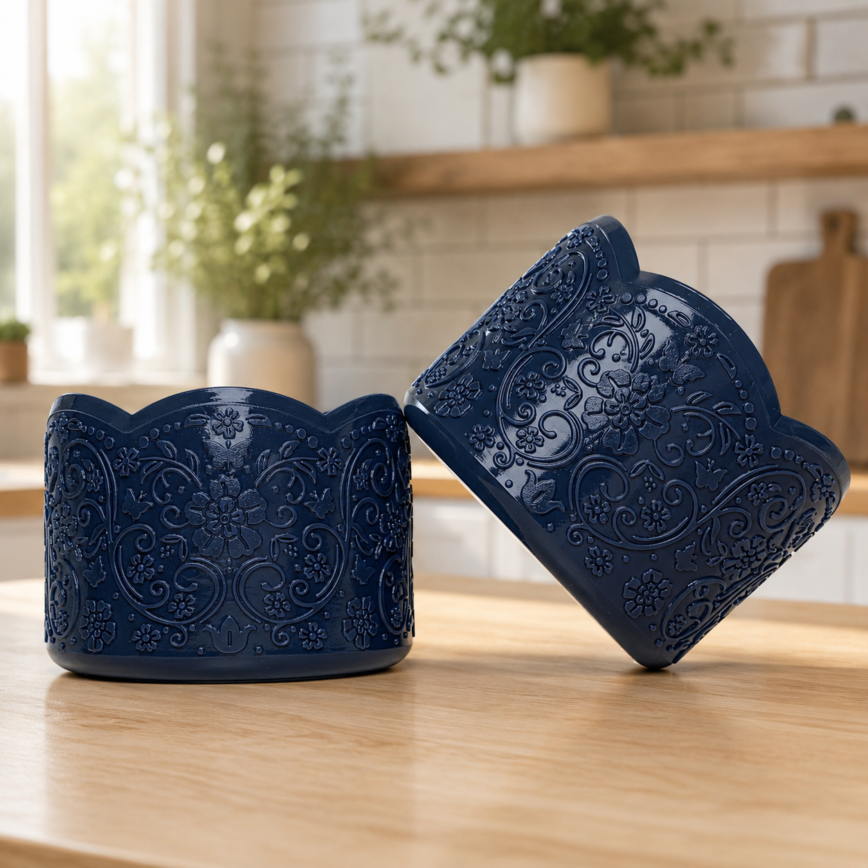 Two shiny navy blue silicone tumbler boots with embossed floral and scroll patterns and scalloped edges, displayed on a light wood table in a bright kitchen setting.
